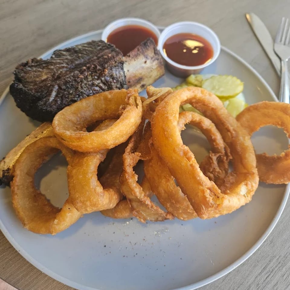 Smokey Sensations BBQ plate with a large rib, onion rings, pickles, and two dipping sauces.