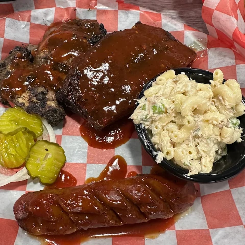 Smokey Sensations BBQ ribs, sausage, macaroni salad, and pickles on red and white checkered paper.