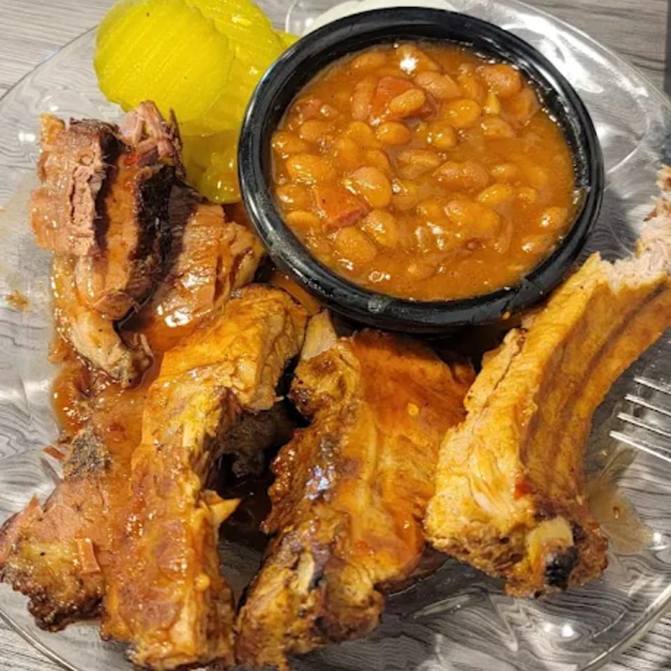 Smokey Sensations BBQ ribs with baked beans and pickles on a clear plate.